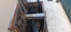 Caixa de esgoto aberta no piso, com a tubulação e o interior enferrujado à mostra