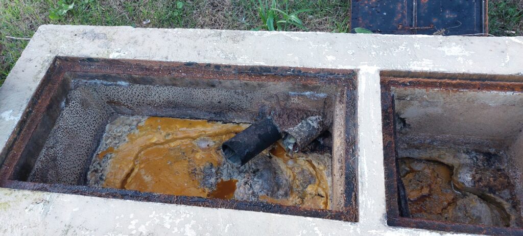 Caixa metálica de esgoto aberta, com resíduo escuro e marcas de ferrugem nas bordas