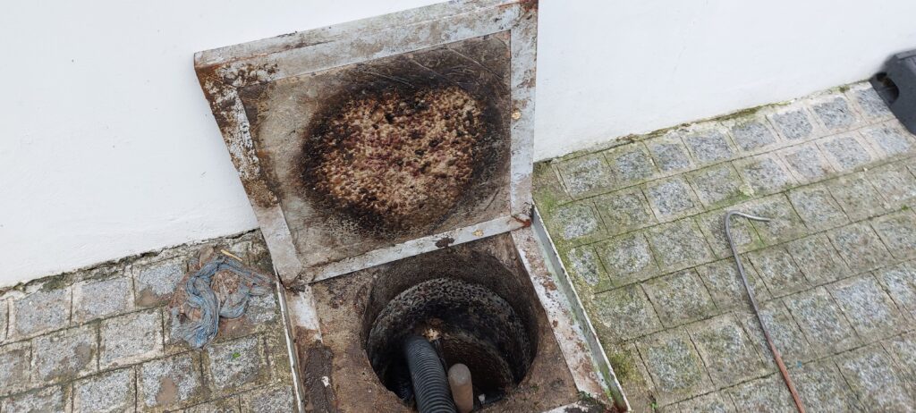 Caixa de esgoto com a tampa aberta ao lado de uma calçada, com uma camada escura de resíduos sobre a abertura