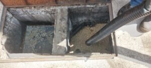 Caixa de esgoto de concreto com sedimento acumulado e uma mangueira de limpeza ao lado