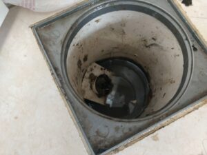 Ralo quadrado do piso visto de cima, com o interior escuro e sujeira acumulada