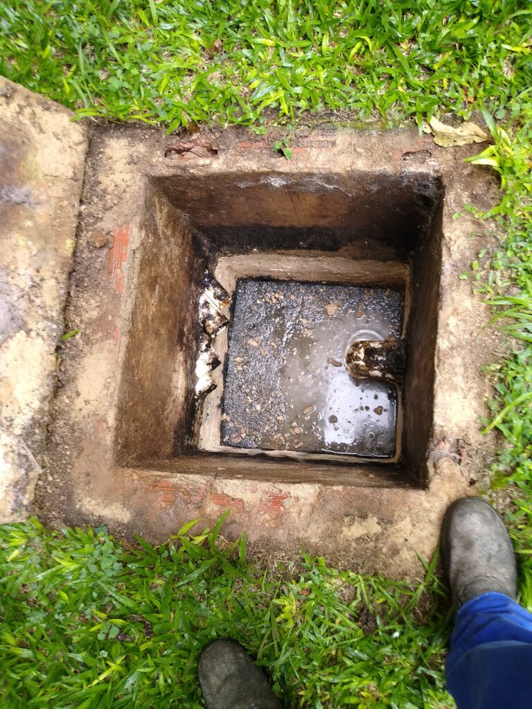 Caixa de esgoto com a tampa aberta em um gramado, com uma grelha e água escura no interior