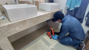Técnico ajoelhado no chão do banheiro, ao lado de uma bancada com duas cubas, operando uma máquina de desentupimento