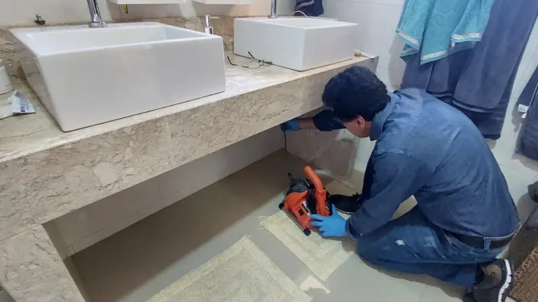Técnico ajoelhado no chão do banheiro, ao lado de uma bancada com duas cubas, operando uma máquina de desentupimento