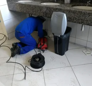 Técnico de desentupidora trabalhando com máquina sob a bancada de um banheiro